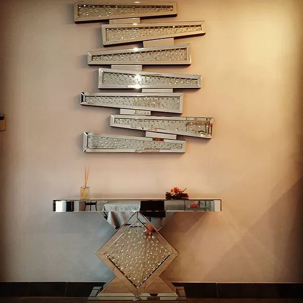 A stylish and modern interior design featuring a mirrored console table with a geometric base and a decorative wall piece composed of several staggered, horizontal, reflective strips, creating a unique visual effect. The setting appears to be a chic, contemporary space.