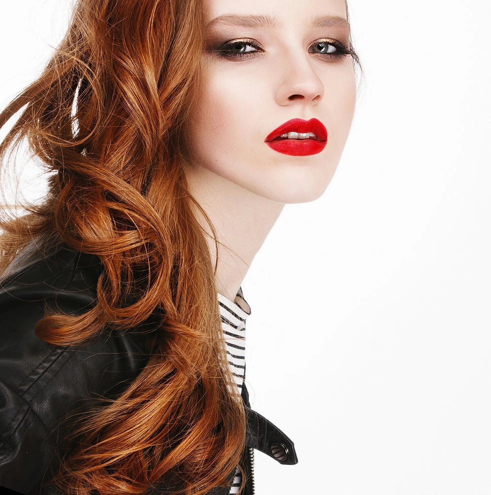 A woman with long red hair, wearing bold makeup with red lipstick, dressed in a black leather jacket, against a plain white background